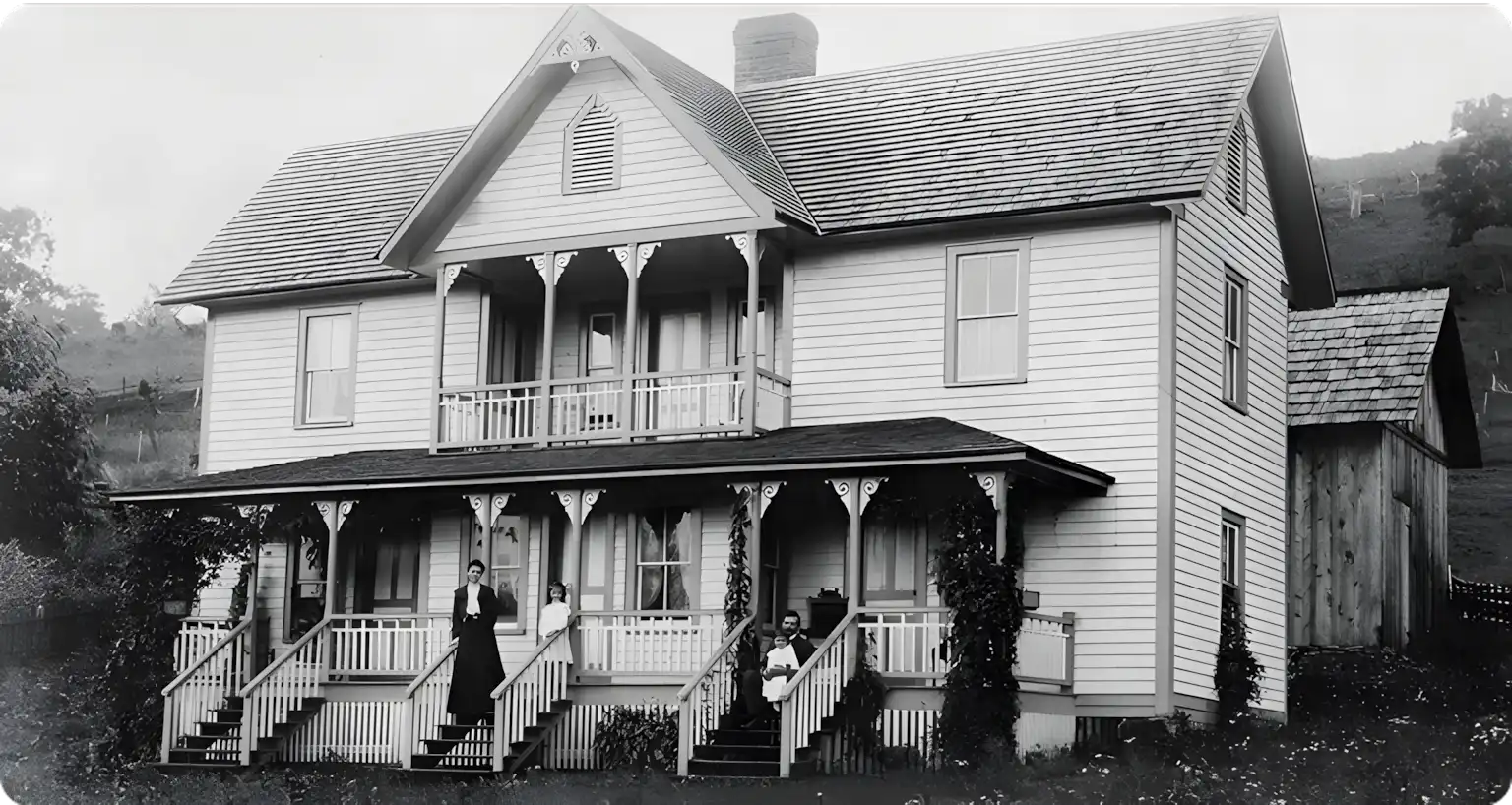 A black-and-white photo of a two-story house with a front porch and balcony, showing three people standing on the steps and porch, with trees and hills in the background.