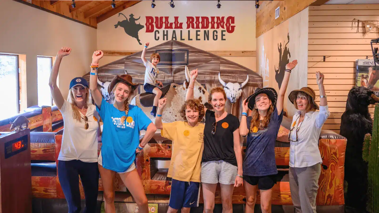 A group of six smiling people in casual clothes and hats pose with raised arms in front of a mechanical bull at an indoor Bull Riding Challenge attraction. A child is riding the bull in the background.