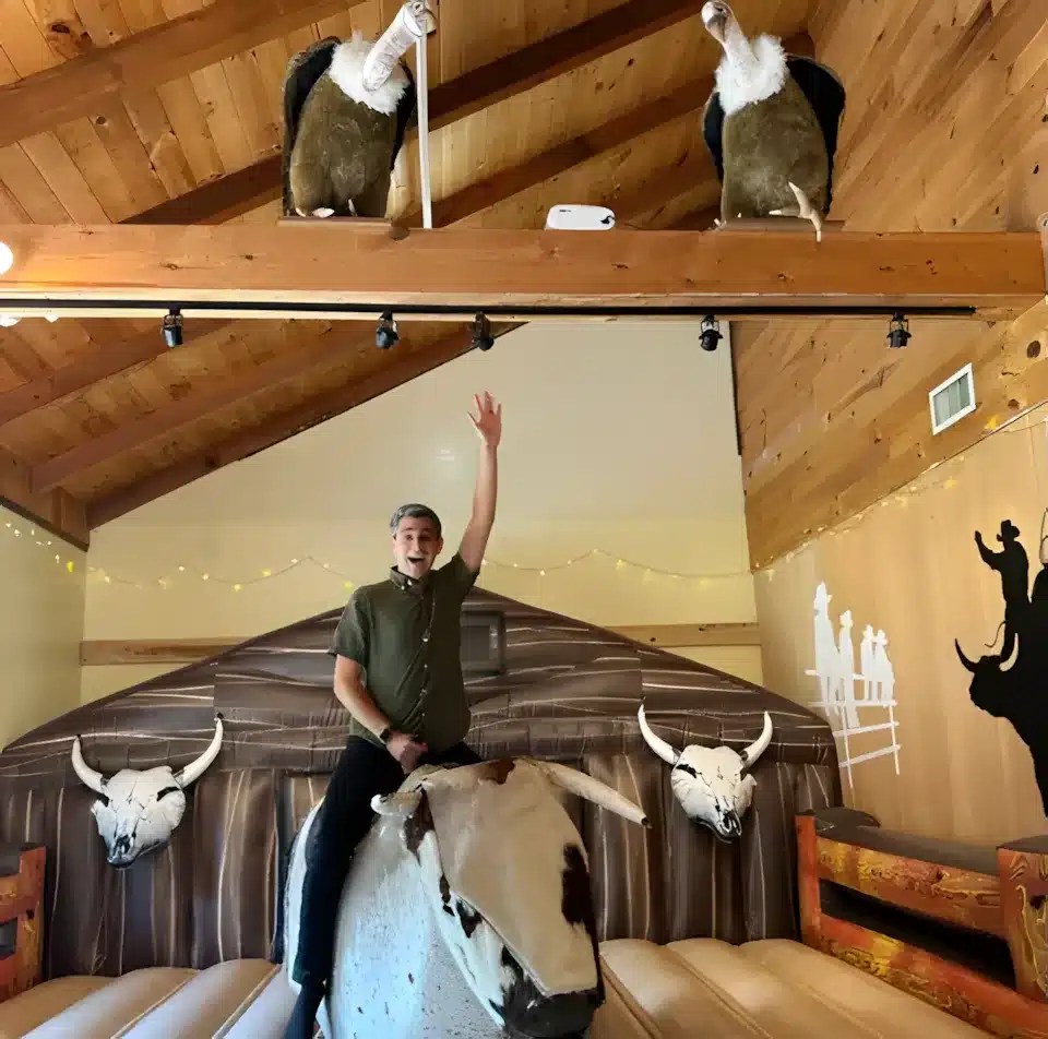 A person with one arm raised rides a mechanical bull indoors. The room has wooden walls and ceiling, with mounted steer skulls and two vulture props perched on a beam above. Western-themed wall art is visible in the background.