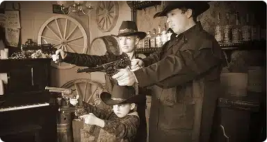 Three people dressed in Old West costumes, including cowboy hats and long coats, pose with toy guns in a vintage-style saloon setting with barrels, bottles, and a wagon wheel in the background.
