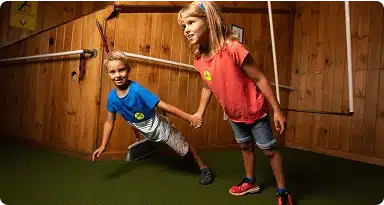 Two children holding hands appear to lean at an extreme angle in a tilted room with wooden walls and green carpet, creating an optical illusion.
