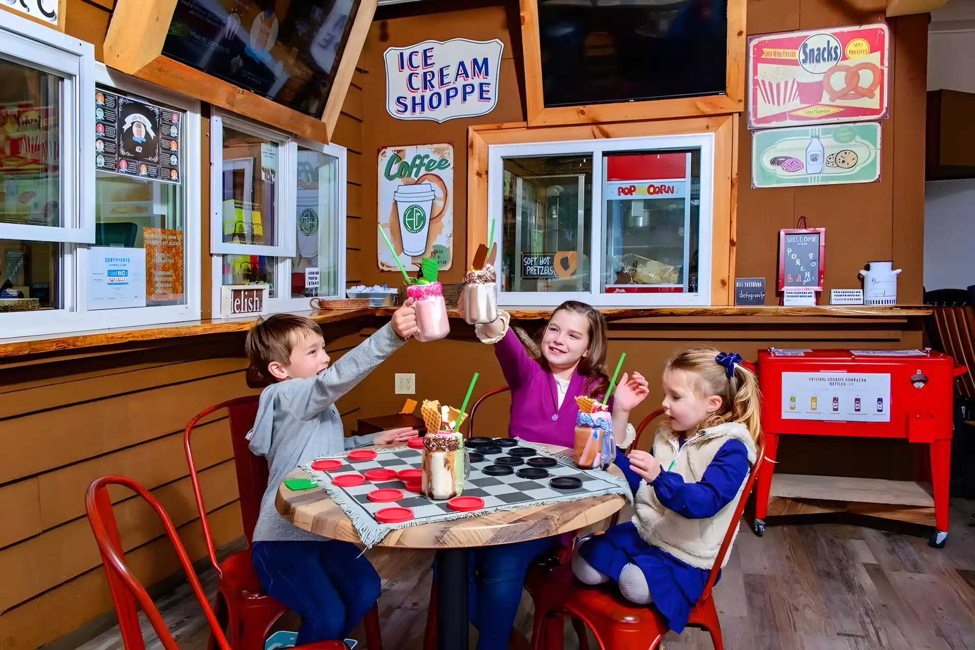 Three young children sit around a table in an ice cream shop, smiling and raising milkshakes in a toast. The shop has colorful signs, wooden walls, and retro décor. A large checkerboard is on the table.