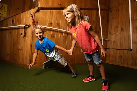 Two children, a boy and a girl, hold hands and lean at an angle in a tilted wooden room, creating an illusion that defies gravity. Both are smiling and appear to be enjoying the playful setting.