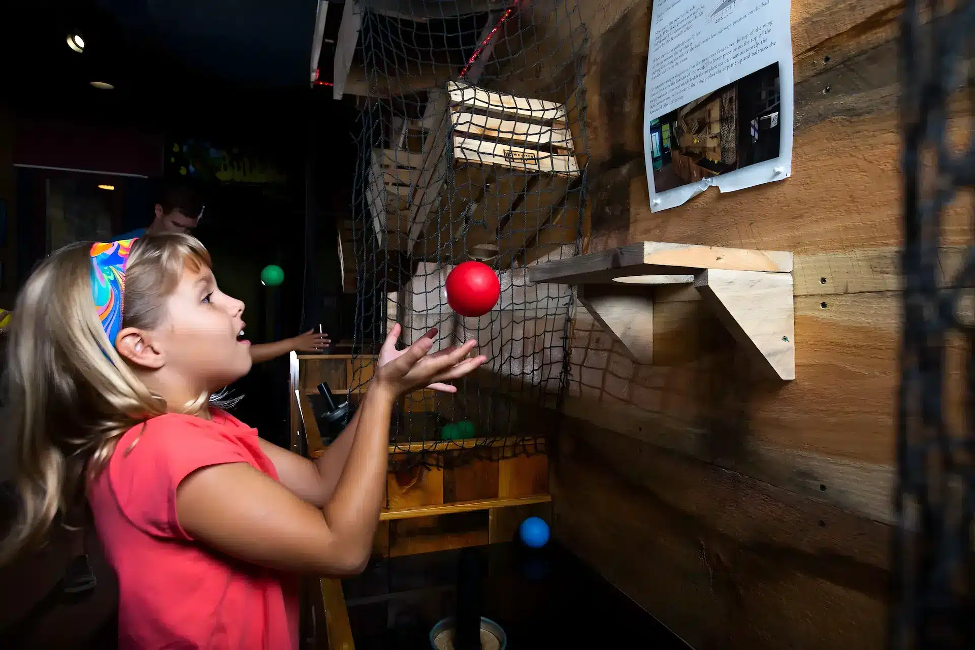 A young girl wearing a headband and red shirt tosses a red ball upwards at an interactive wooden exhibit, with wooden ramps and a net on the wall behind her.
