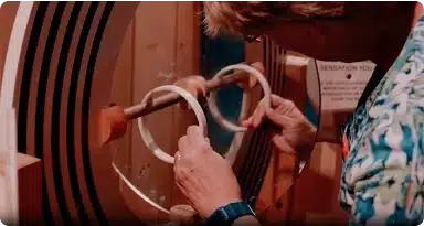 A person wearing glasses and a patterned shirt holds two circular wooden rings, positioning them on a spindle inside a woodworking machine.