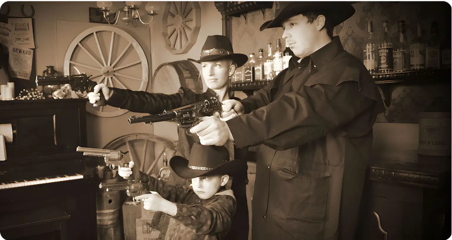 Three people dressed in cowboy outfits and hats pose with toy guns in an old-fashioned Western-style saloon setting, featuring wooden barrels, wagon wheels, and bottles on shelves in the background.