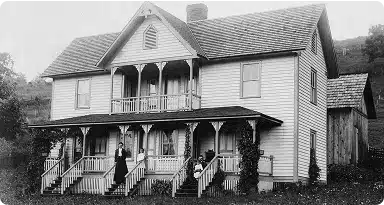 A black-and-white photo of a two-story house with a covered front porch and balcony. Three people stand or sit on the porch, and vines grow up the columns. The house is surrounded by grass and trees.