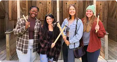 Four women stand side by side smiling and holding axes, posing in front of wooden targets at an outdoor axe-throwing venue.