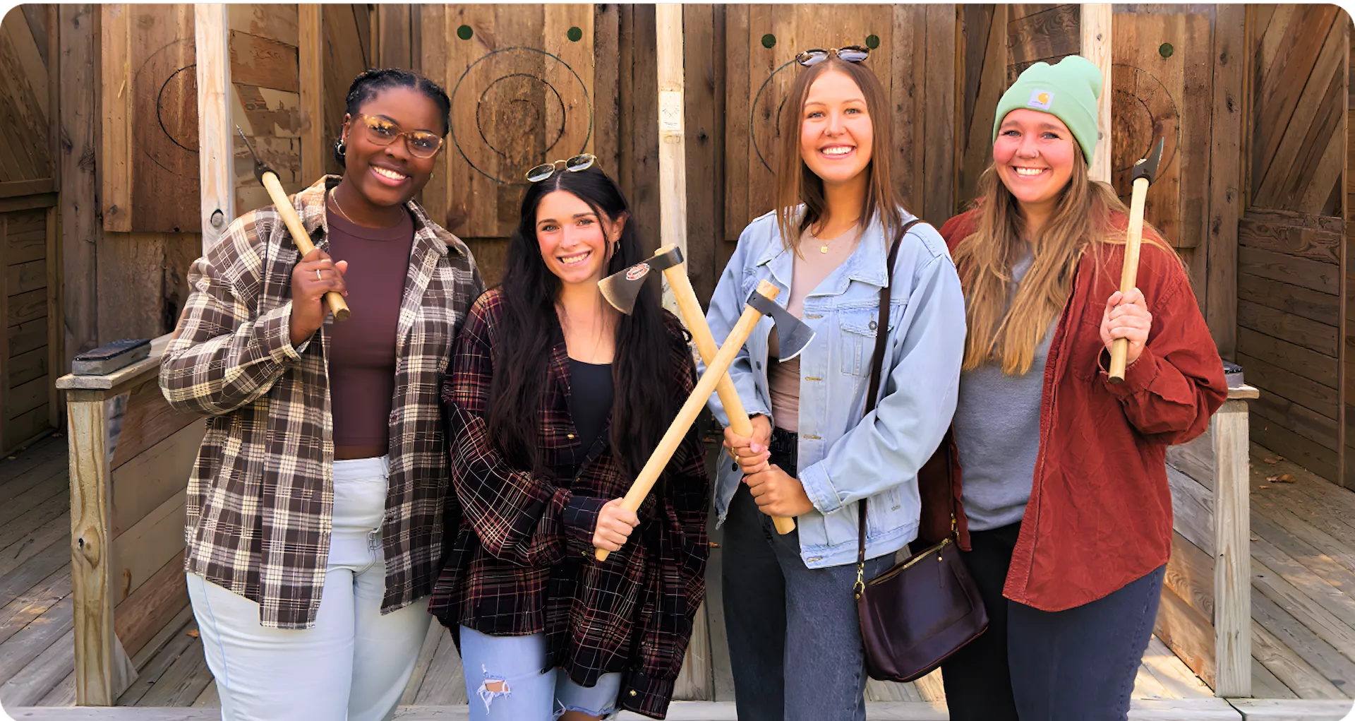 Four smiling women stand side by side outdoors, each holding a throwing axe, in front of wooden targets. They are dressed casually in jackets, flannel shirts, and jeans.