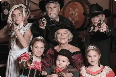 A family dressed in old-fashioned Western costumes poses for a photo. The adults and children wear hats, bonnets, and dresses, and some hold toy guns, creating a playful Wild West scene.
