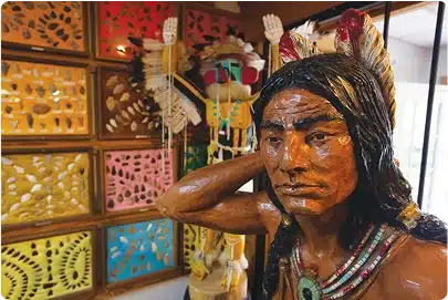 A wooden statue of a Native American man with long hair and a feather headdress stands in front of colorful displays of arrowheads and a traditional Native American figure in the background.