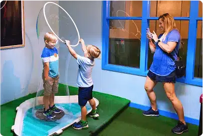 Two young boys play with a large bubble wand, surrounding one boy with a bubble, while a woman in blue takes a photo and smiles in a brightly lit indoor play area with green flooring and blue windows.