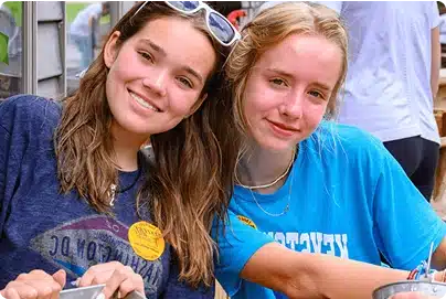 Two young women smile at the camera while sitting close together outdoors. They are wearing casual t-shirts, and one has sunglasses on her head. Both appear relaxed and happy.