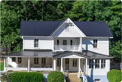 White two-story farmhouse with a black metal roof, wraparound porch, upper balcony, and steps leading to the front entrance, surrounded by green bushes and trees in the background.