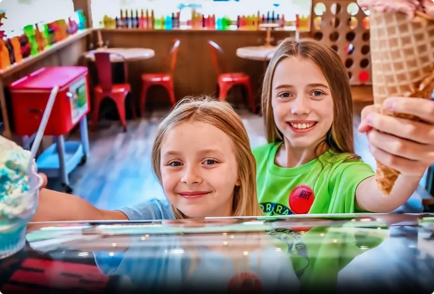Two smiling children stand at an ice cream shop counter, holding up colorful ice cream cones and a cup. The background shows bright chairs, tables, and bottles of toppings on a shelf.