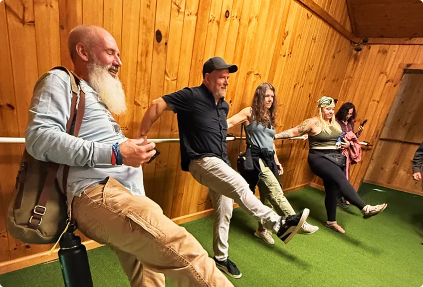 Five adults stand in a row inside a wooden room, lifting one leg and smiling as they balance together, appearing to participate in a fun group activity.