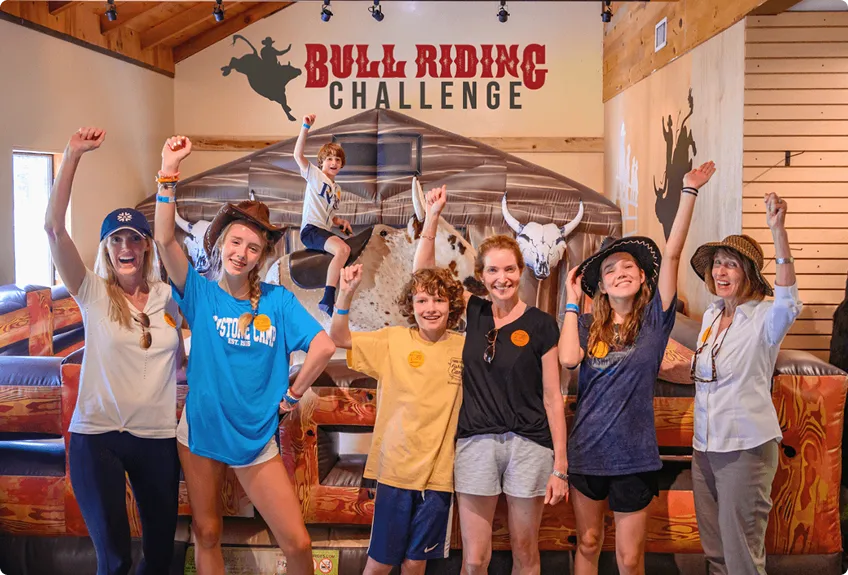 A group of six smiling people pose with raised arms in front of a mechanical bull ride at an indoor Bull Riding Challenge venue. A child sits on the bull in the background, also raising an arm.