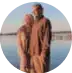Two people stand close together by a body of water, both wearing light brown clothing. The background shows a calm, blue sky and water, suggesting a peaceful outdoor setting.