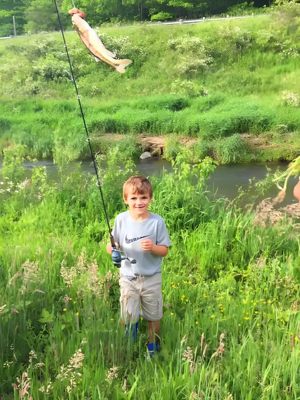A young boy stands in tall grass by a stream, smiling and holding a fishing rod with a large fish dangling from the line. Lush greenery and wildflowers surround him.