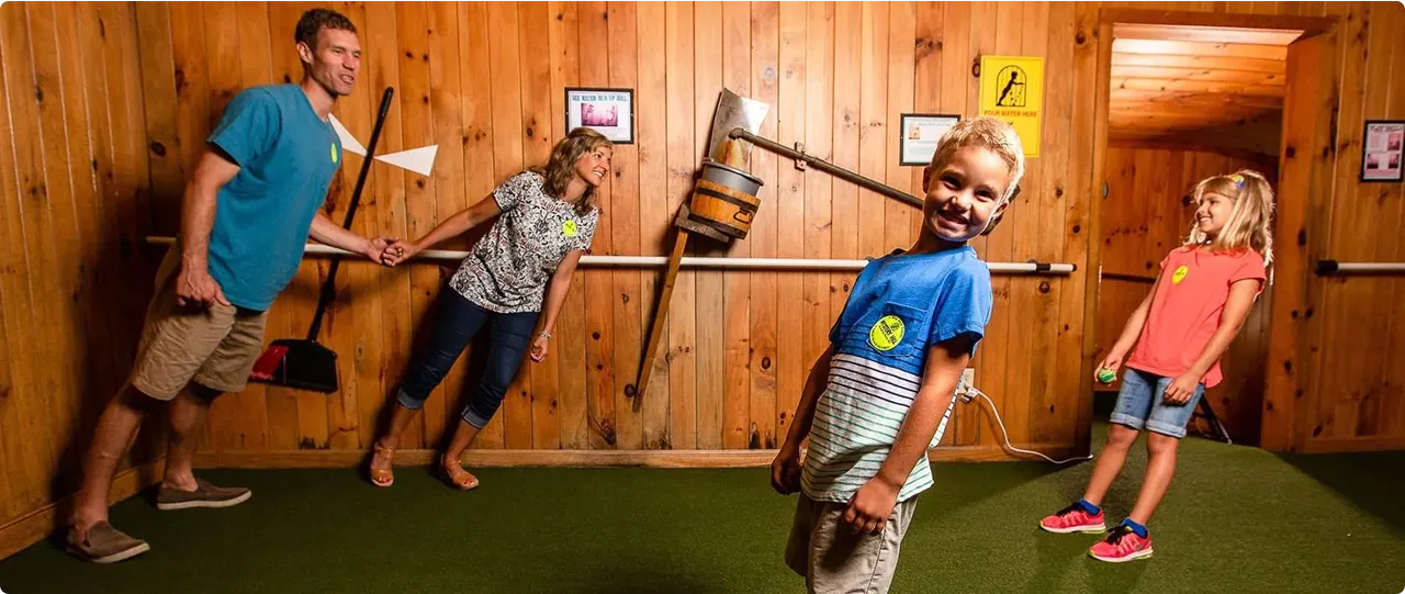 A family of four inside a wooden room with slanted walls, creating the illusion that they are leaning at odd angles. They are smiling and appear to be enjoying the quirky experience.