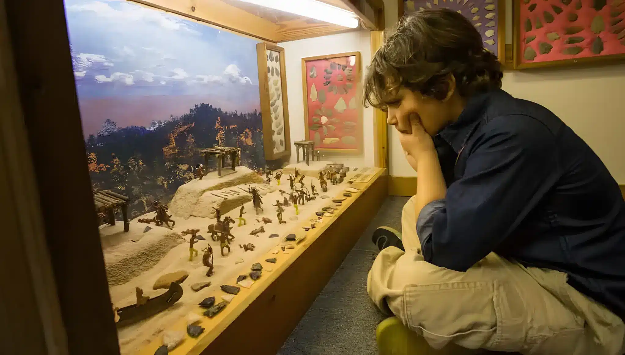 A boy crouches and looks thoughtfully at a detailed miniature diorama of a village scene with tiny figures and buildings, displayed in a glass case at a museum.