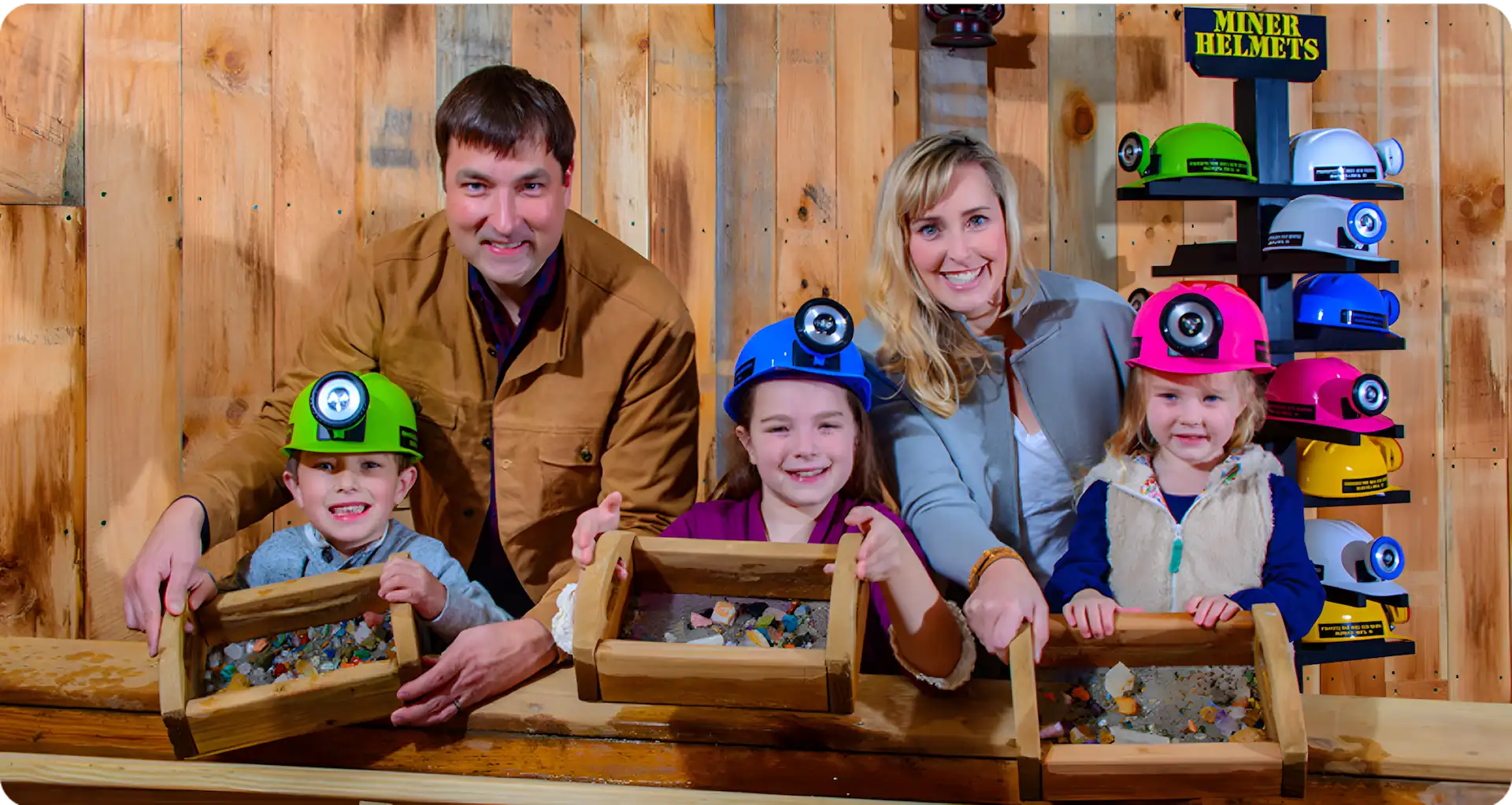 A smiling family of four, two adults and two children, pose indoors with mining helmets and wooden trays filled with rocks, standing at a wooden table near a rack displaying colorful miner helmets.