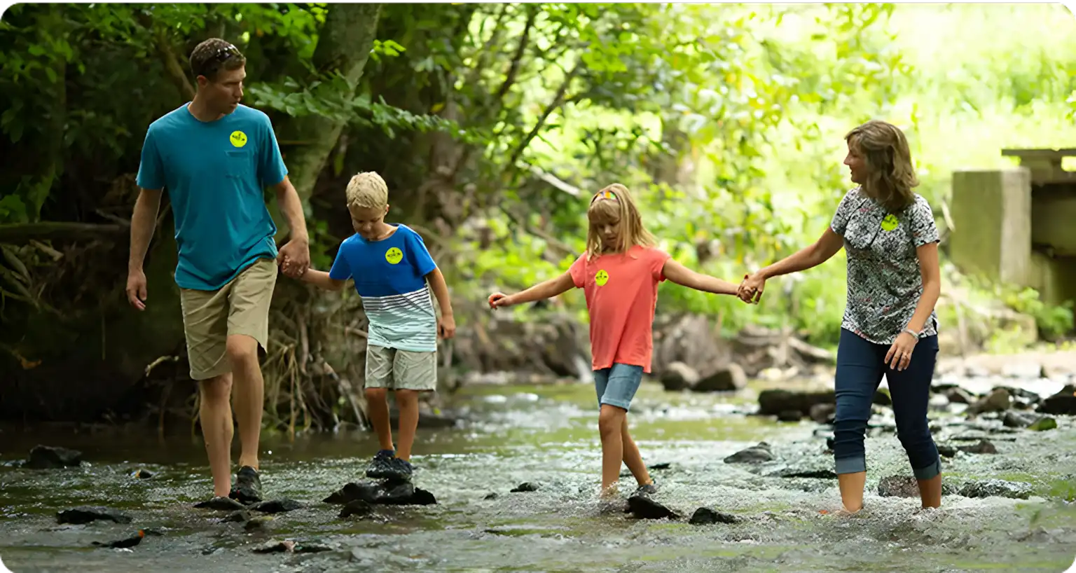 A family of four, two adults and two children, walk hand in hand across a shallow creek surrounded by lush green trees. The children are in the center, and everyone wears casual summer clothes and yellow badges.