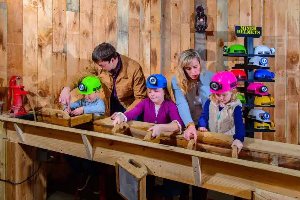 A family with two adults and three children wearing colorful miner helmets sift for gems in wooden troughs indoors; multiple helmets are displayed on shelves behind them.