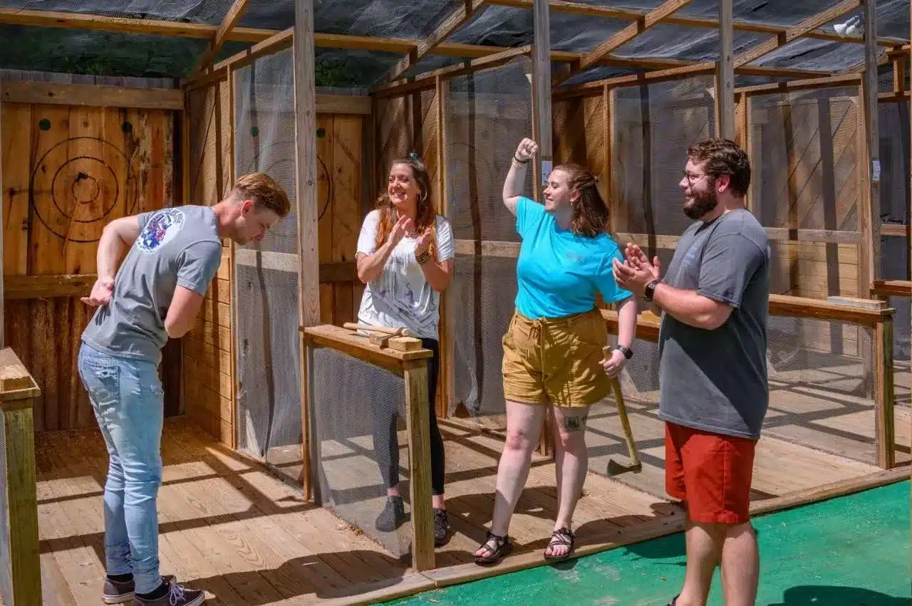 Four people stand in an outdoor axe-throwing lane; one person bows playfully while the others cheer and clap, celebrating a successful throw. The setting is sunny with wood and mesh enclosures in the background.