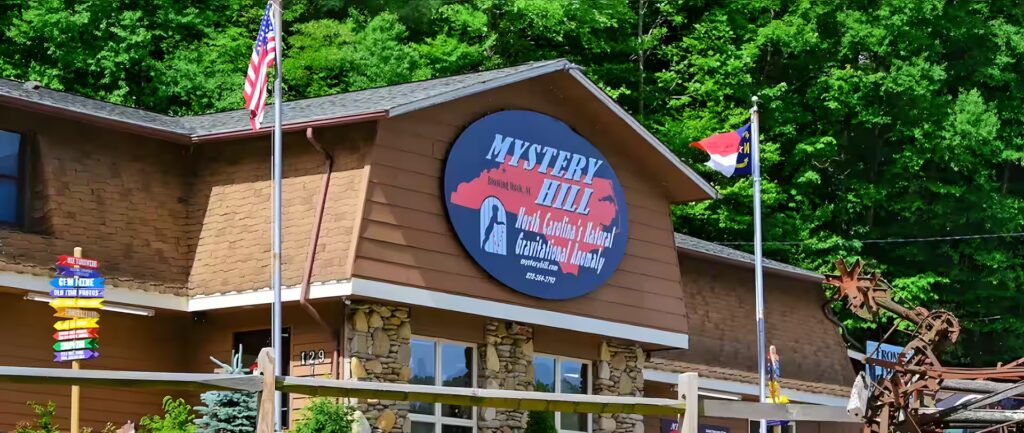 A brown building with a large sign reading Mystery Hill, displaying American and North Carolina flags, surrounded by trees and colorful signposts, under a clear sky.